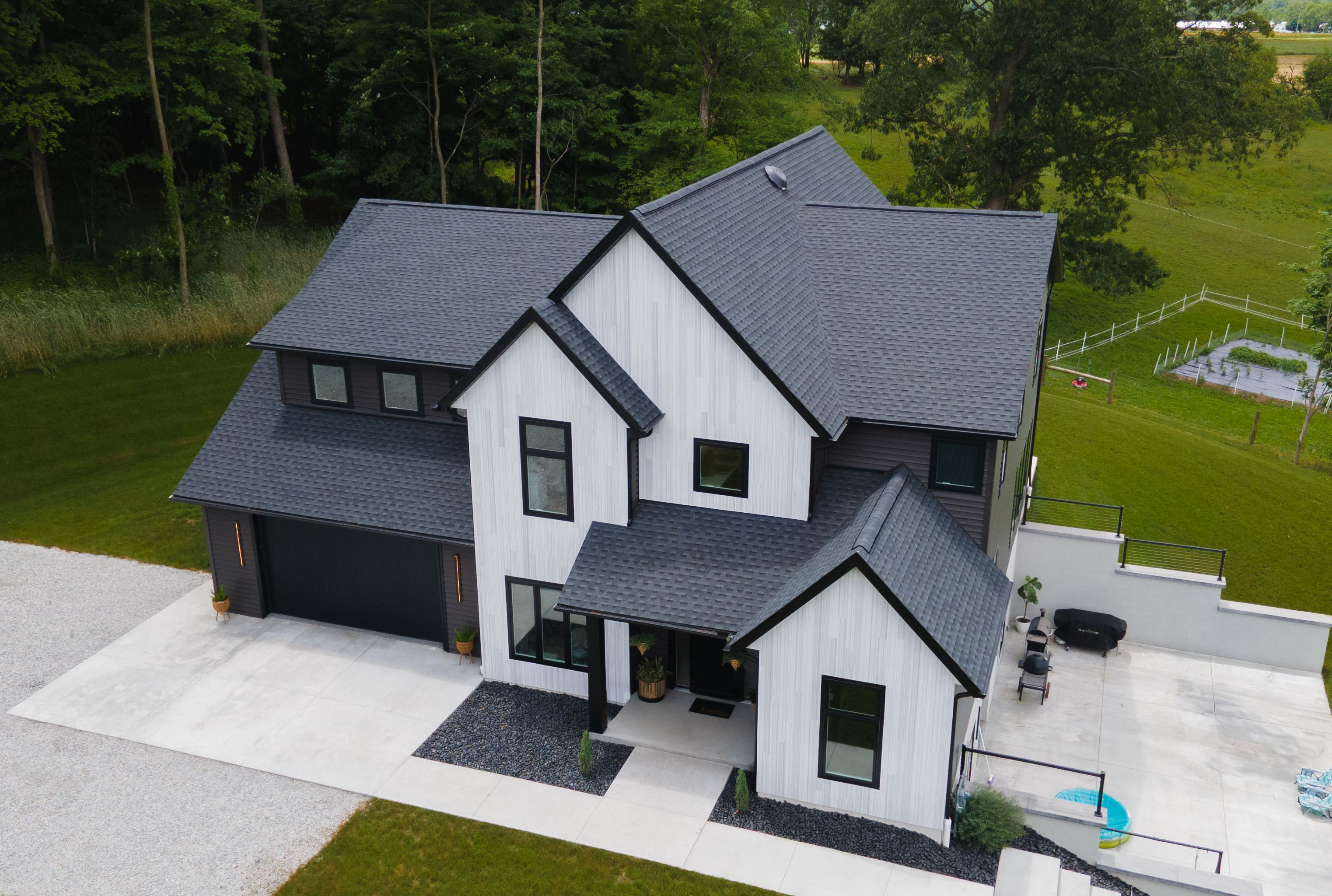 A modern two-story house with gray shingle roofs, black-framed windows, and white exterior walls, surrounded by green grass and trees with a paved driveway.