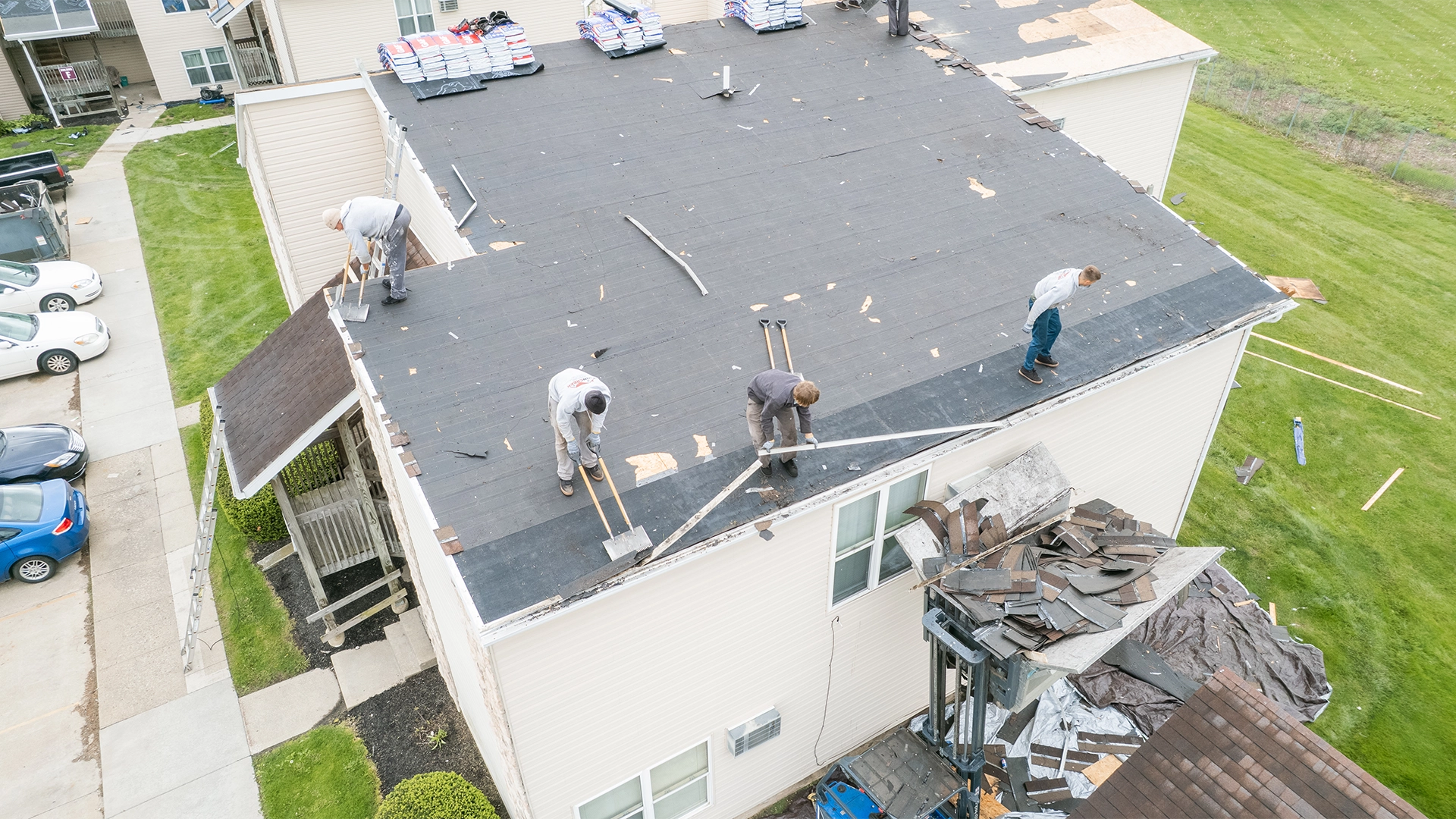 Four workers are replacing a flat roof on a light-colored residential building, handling roofing materials and tools, with removed shingles piled on a lift platform beside the building.