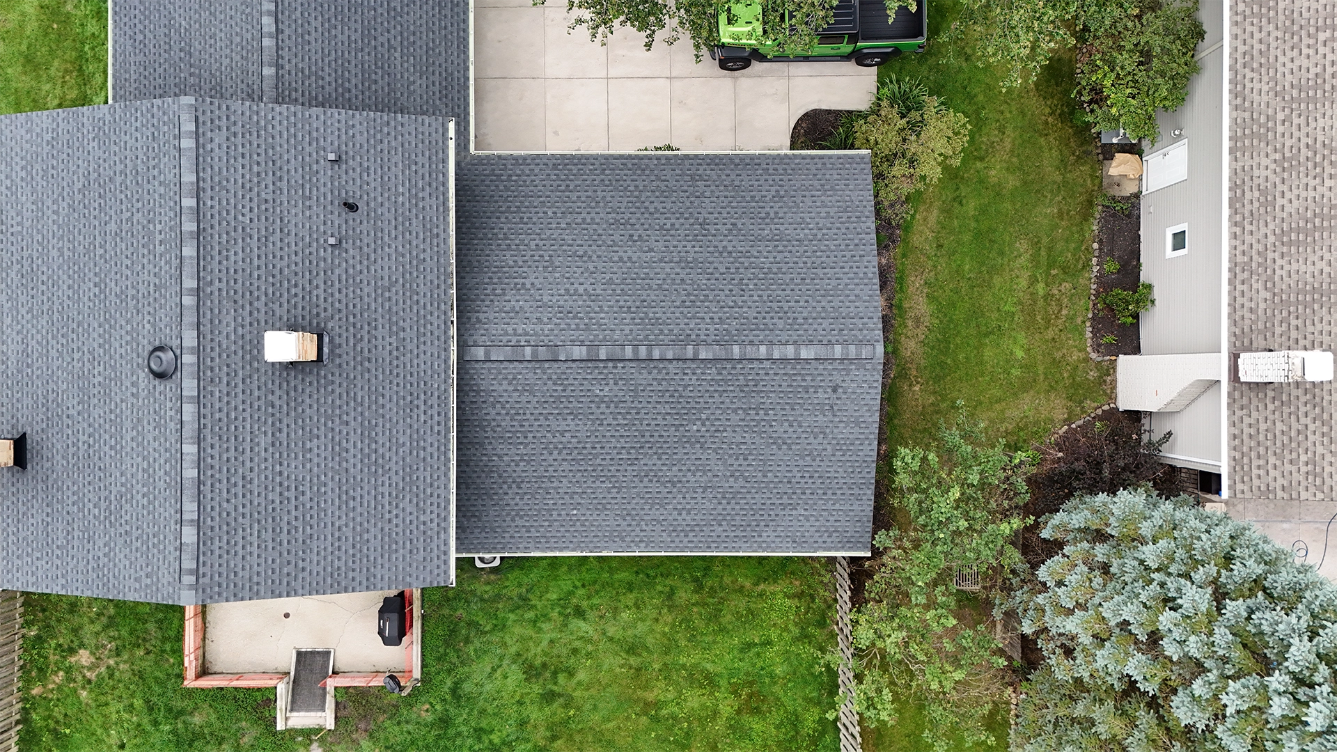 Aerial view of two adjacent houses with gray shingled roofs, green lawns, trees, a green vehicle parked in a driveway, and a concrete patio with a grill.