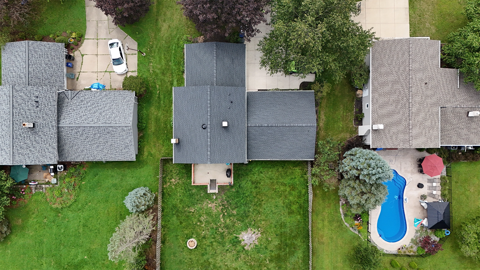 Aerial view of three suburban houses with gray roofs, green lawns, a driveway with a white car, and a fenced backyard with a blue swimming pool and patio furniture under a red umbrella.