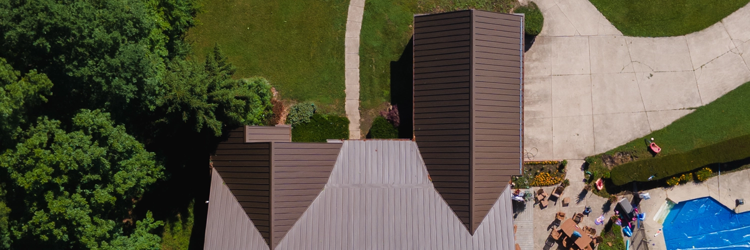 Aerial view of a residential property showing a large brown metal roof surrounded by green trees, a concrete driveway, patio furniture, and a blue swimming pool with a cover partially visible.
