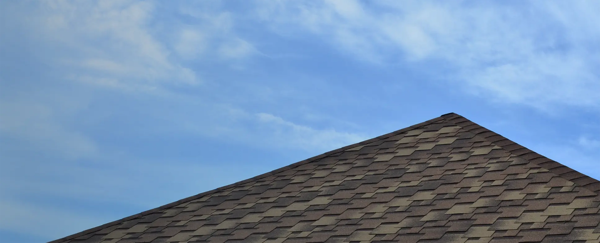 Peak of a brown architectural shingle roof against a blue sky with wispy clouds.