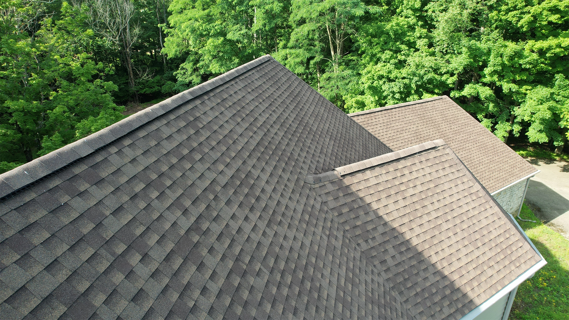 Drone view of a weathered-wood brown architectural shingle roof with multiple gables, surrounded by green trees.
