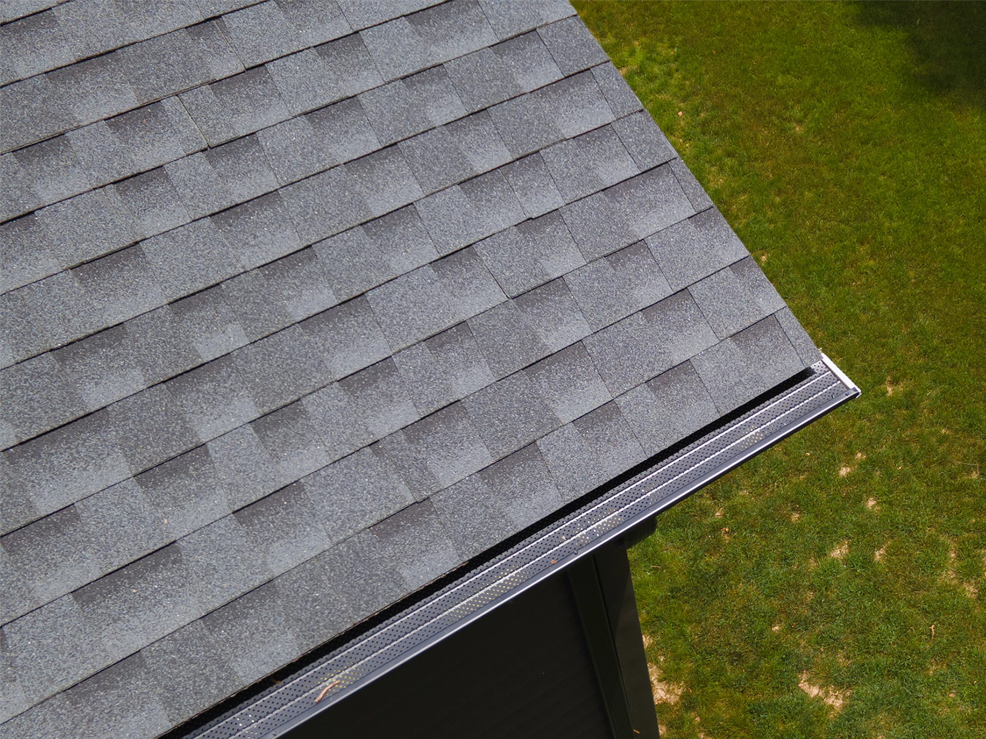 Close-up of a gray asphalt shingle roof edge with a perforated gutter guard installed over a black seamless gutter.