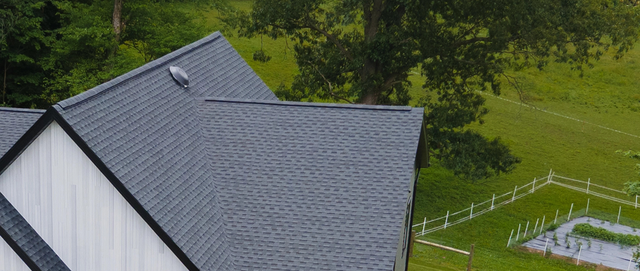 Close-up of a roof with damaged and lifted asphalt shingles under trees in the background.