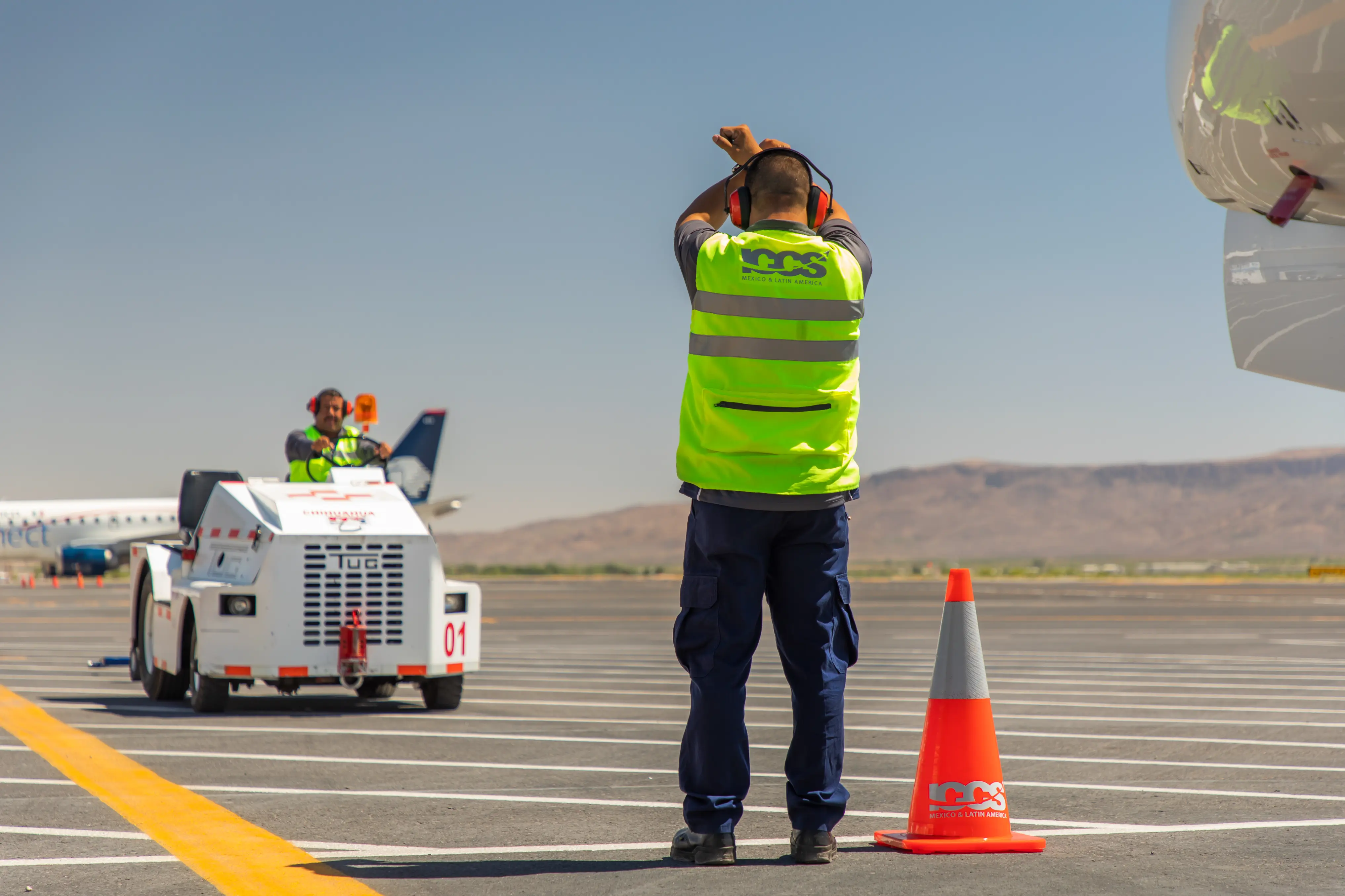 Airport ground staff in high-visibility vests guiding a white aircraft tug on the tarmac under clear skies.