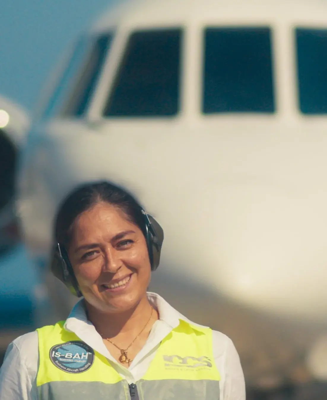 Smiling female ground crew member wearing a yellow safety vest and headphones in front of an airplane cockpit.