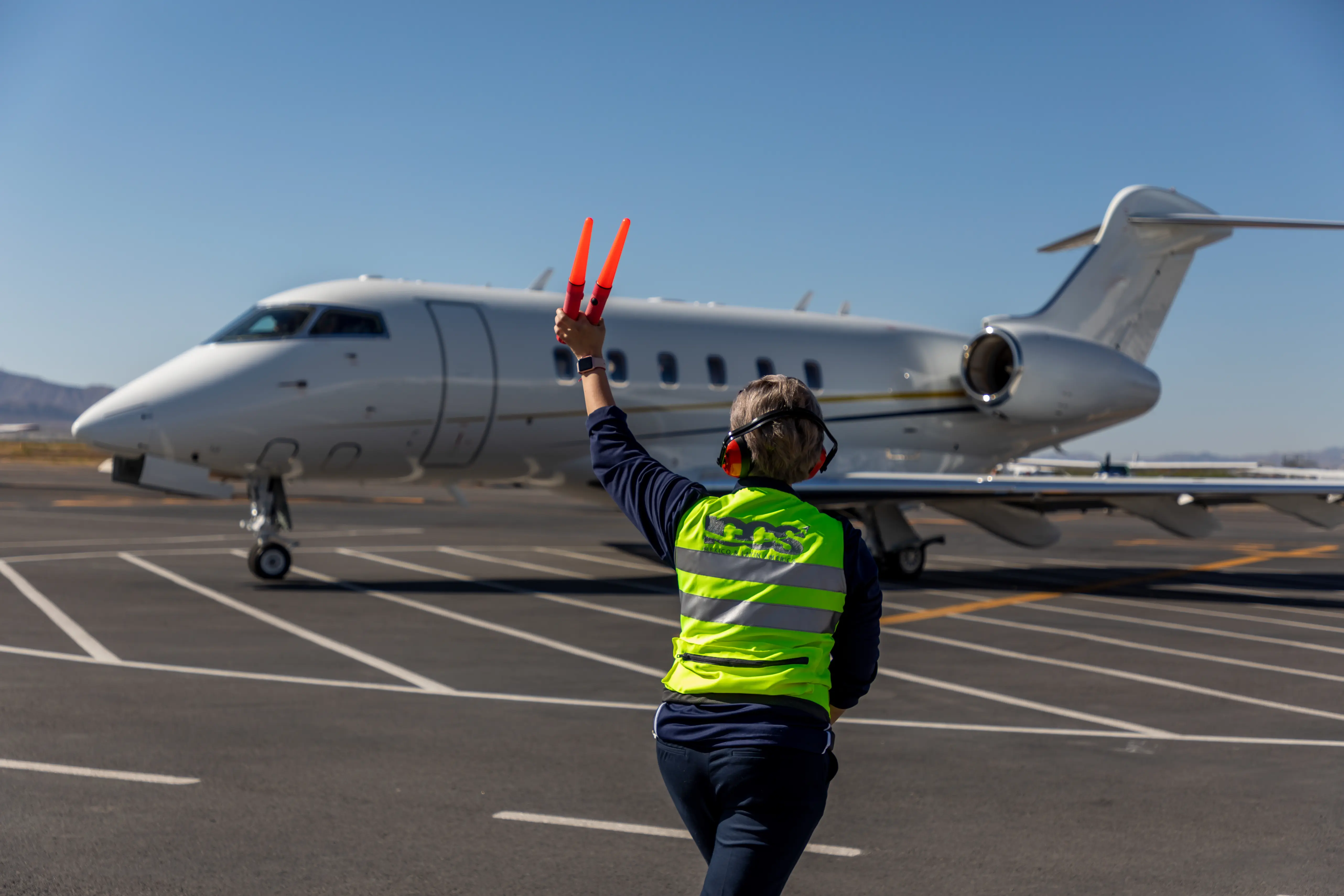 Ground crew member in a neon safety vest and headphones directing a private jet on the tarmac using orange marshalling wands.