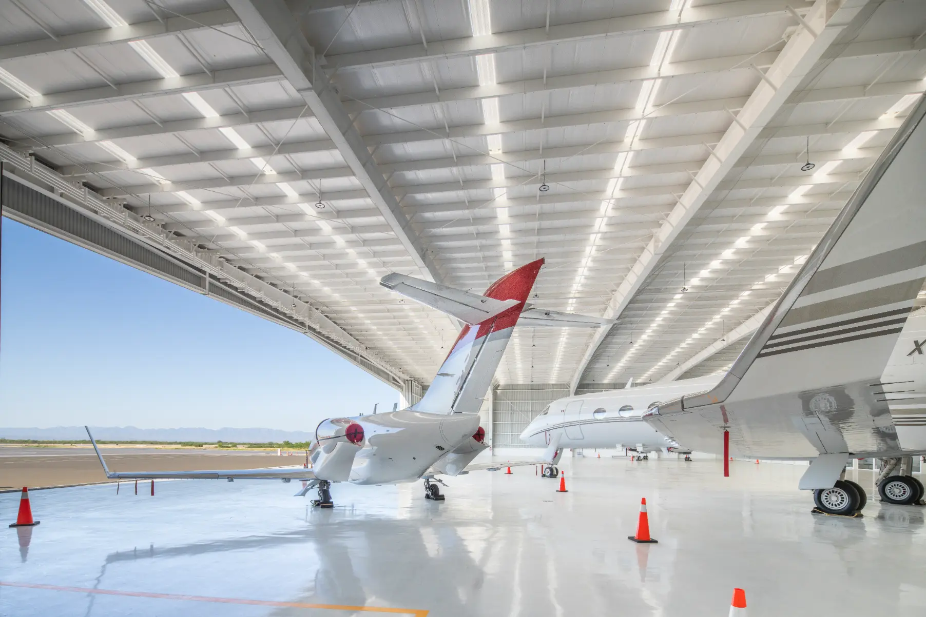 Two private jets parked inside a spacious, brightly lit aircraft hangar with orange safety cones around them.