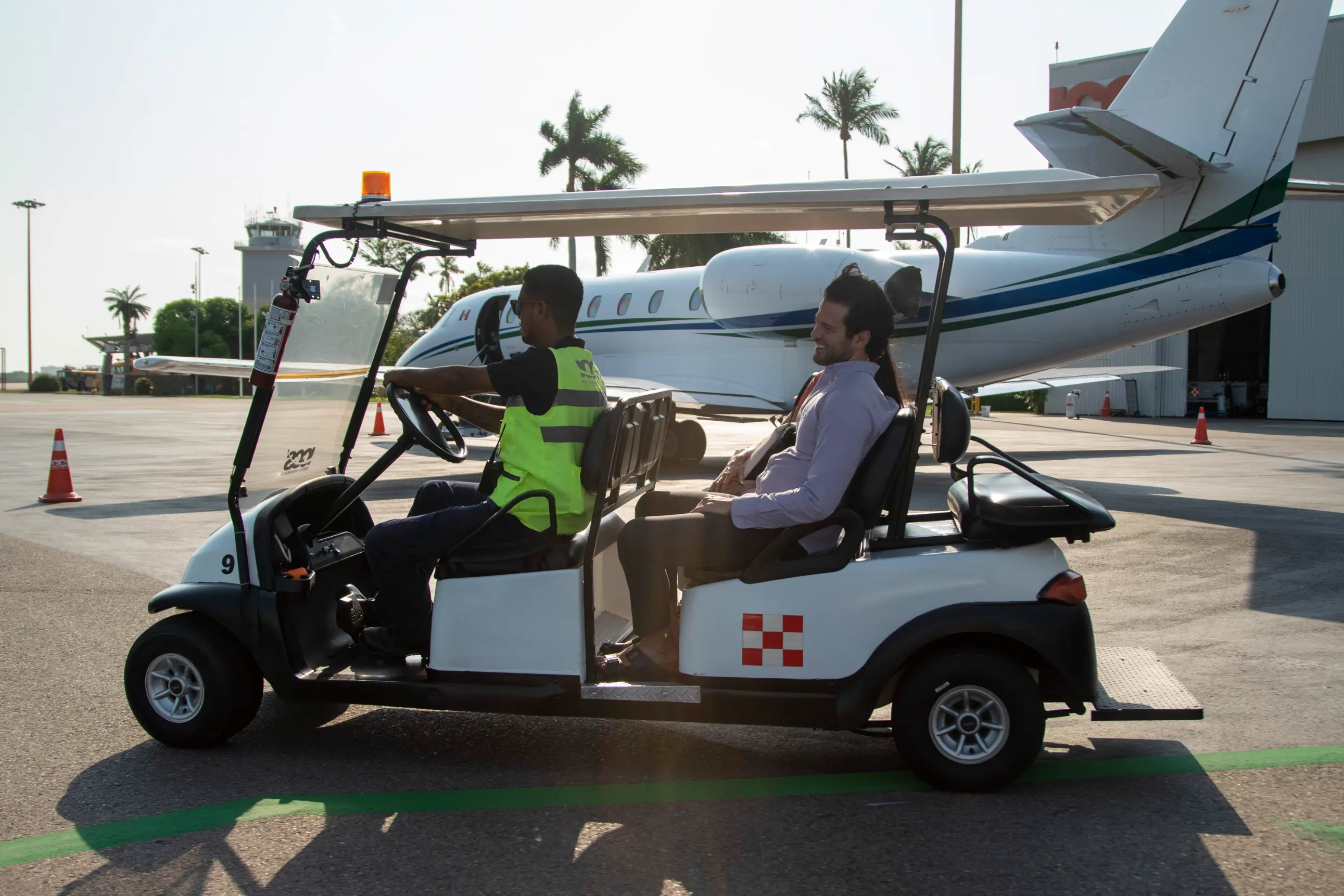 Man wearing a neon safety vest driving a golf cart with a passenger on an airport tarmac near a private jet.