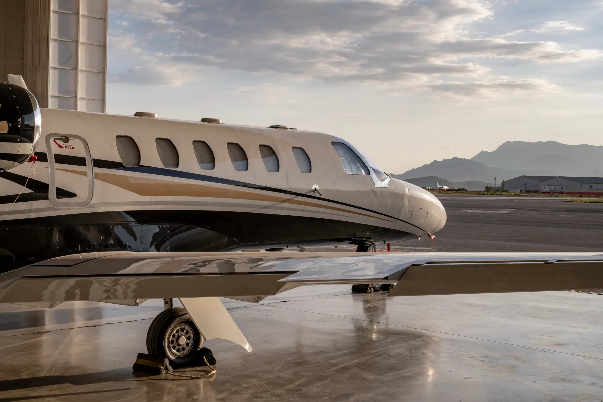 Side view of a private jet on the tarmac with mountains and a hangar in the background under a cloudy sky.