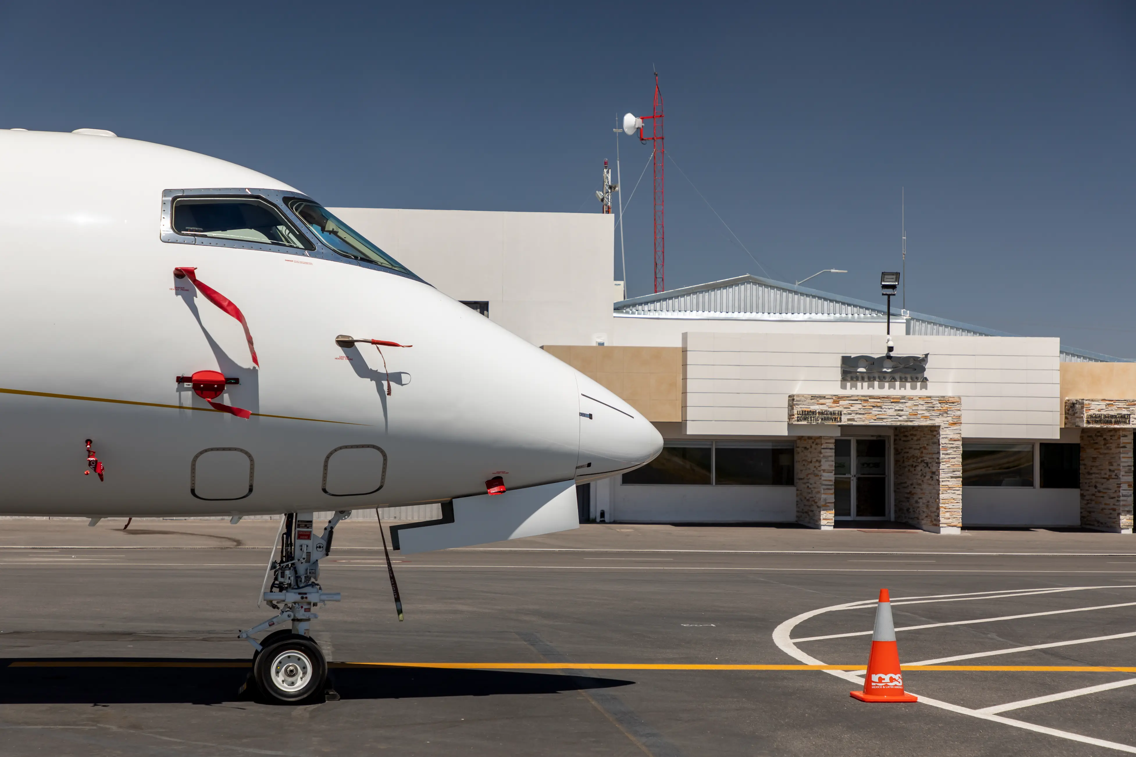 Close-up of a white airplane nose and front landing gear on the tarmac near an airport building with a red communications tower.