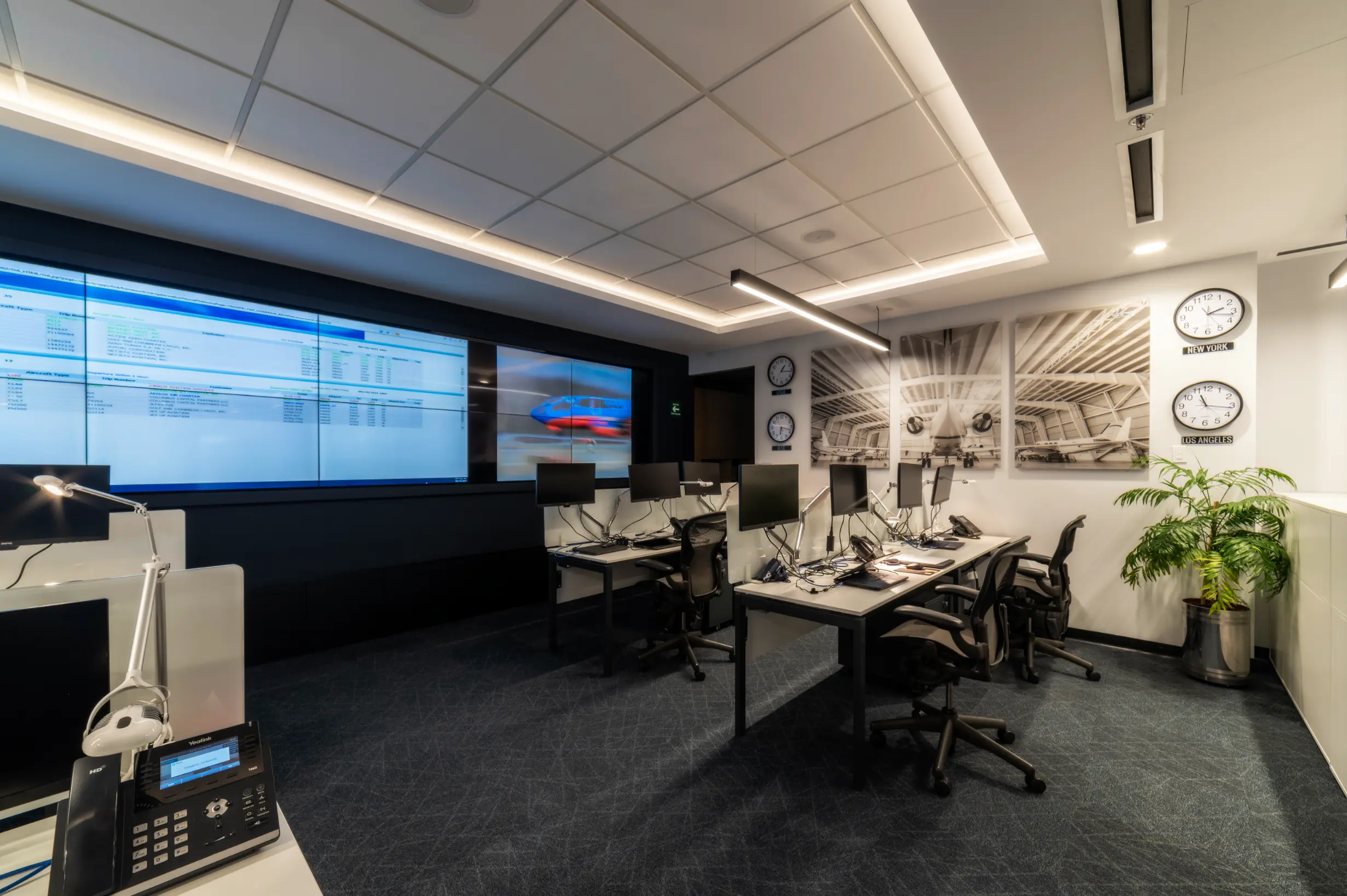 Modern office space with multiple desks, computer monitors, large wall screens displaying data and images, clocks showing different time zones, and aviation-themed wall art.
