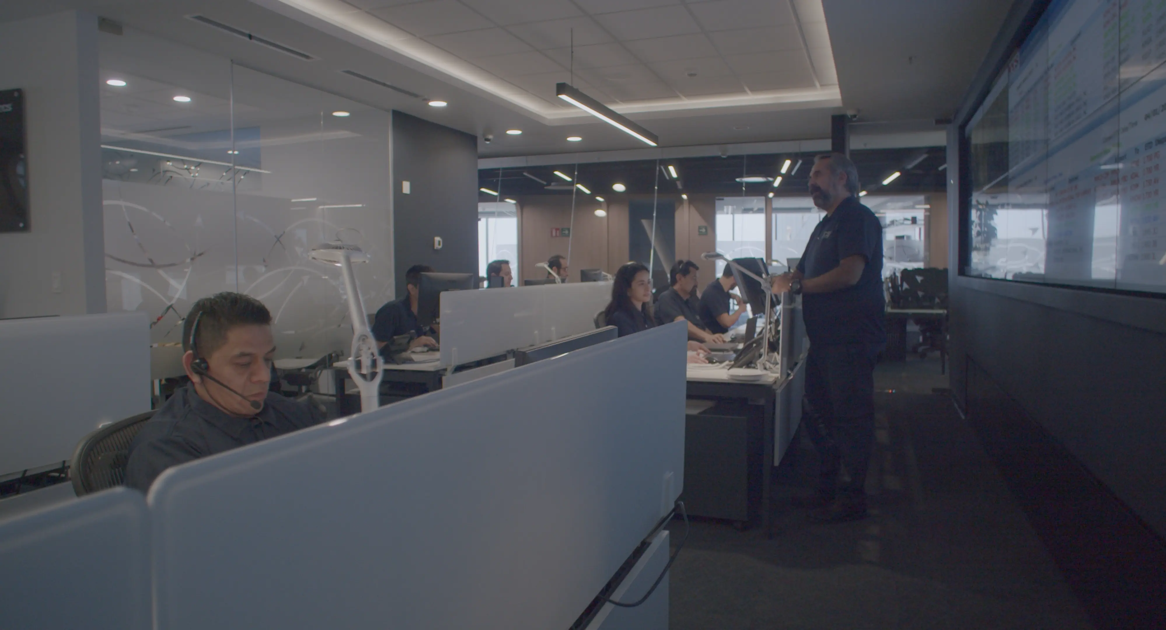 Office workspace with multiple people working at desks and a man standing near a large screen display on the wall.