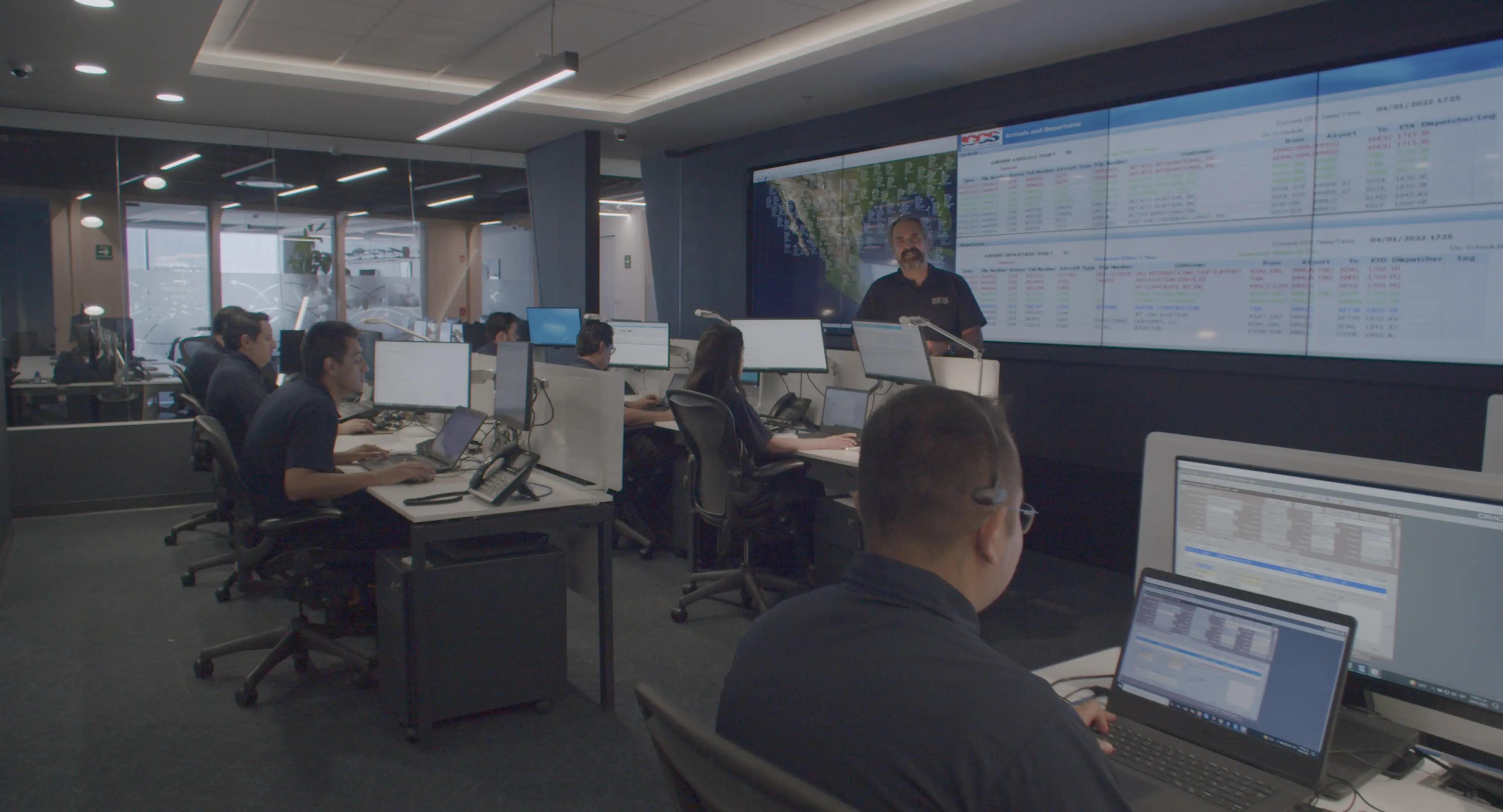 Office workspace with employees at desks working on multiple computer monitors and a man standing in front of a large screen displaying a map and data.