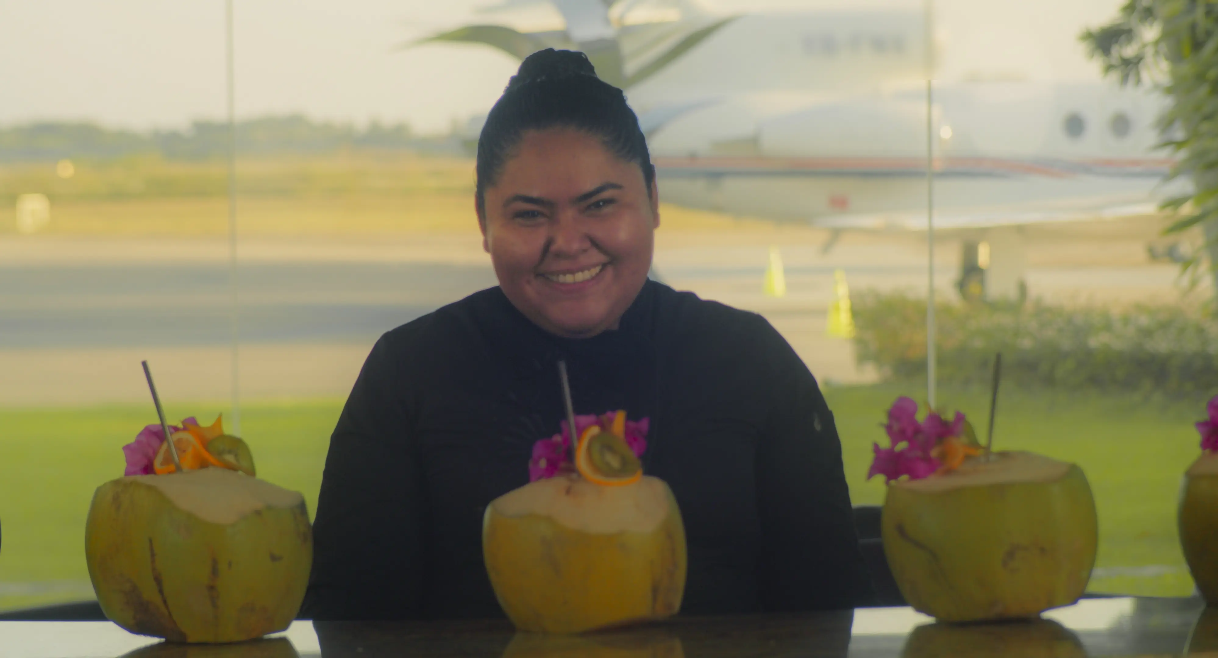 Smiling woman sitting behind a table with three decorated coconuts, with an airplane visible outside through large windows.