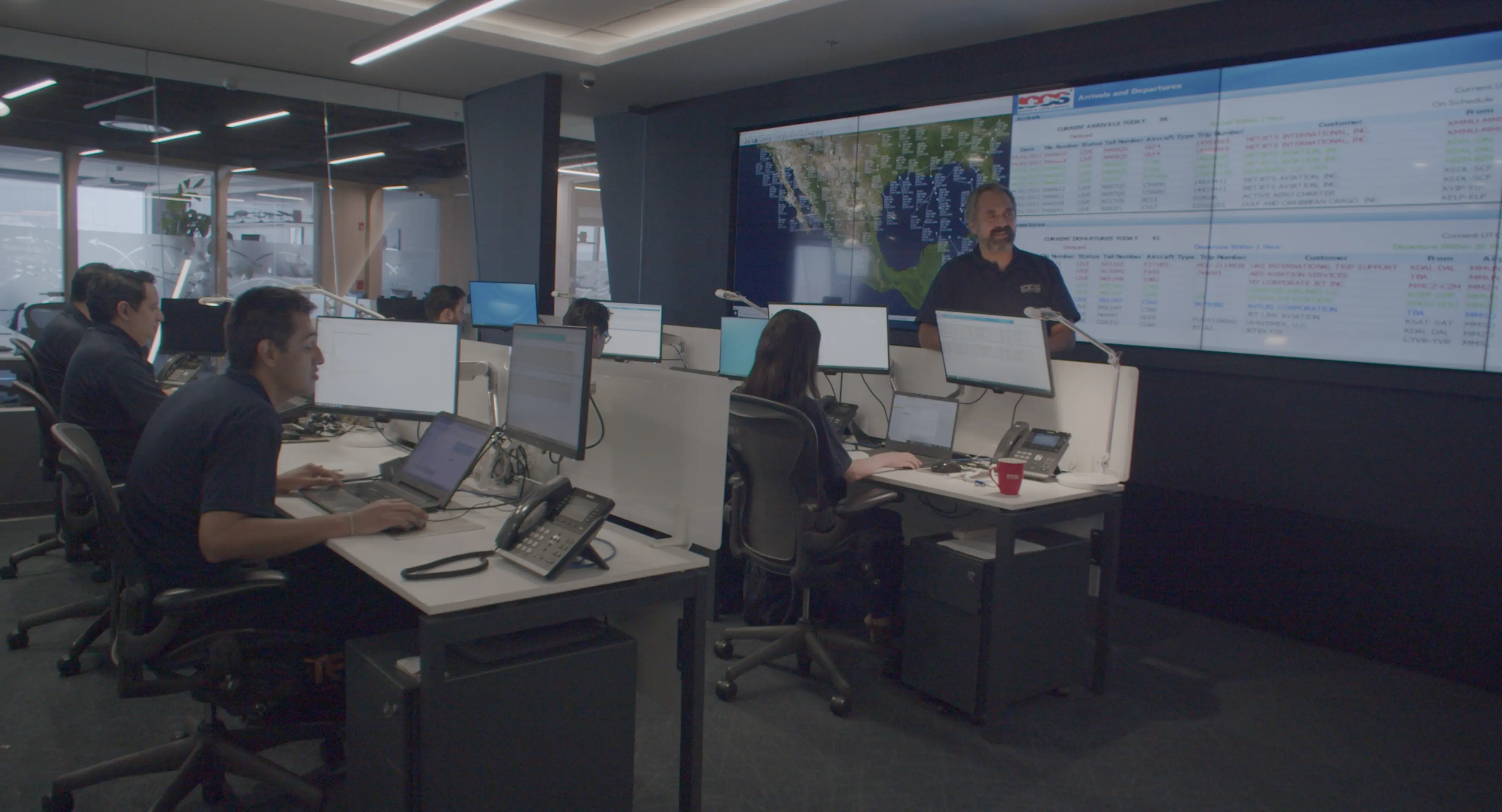 Office setting with people working at desks with multiple monitors and a large screen displaying maps and data on the wall.