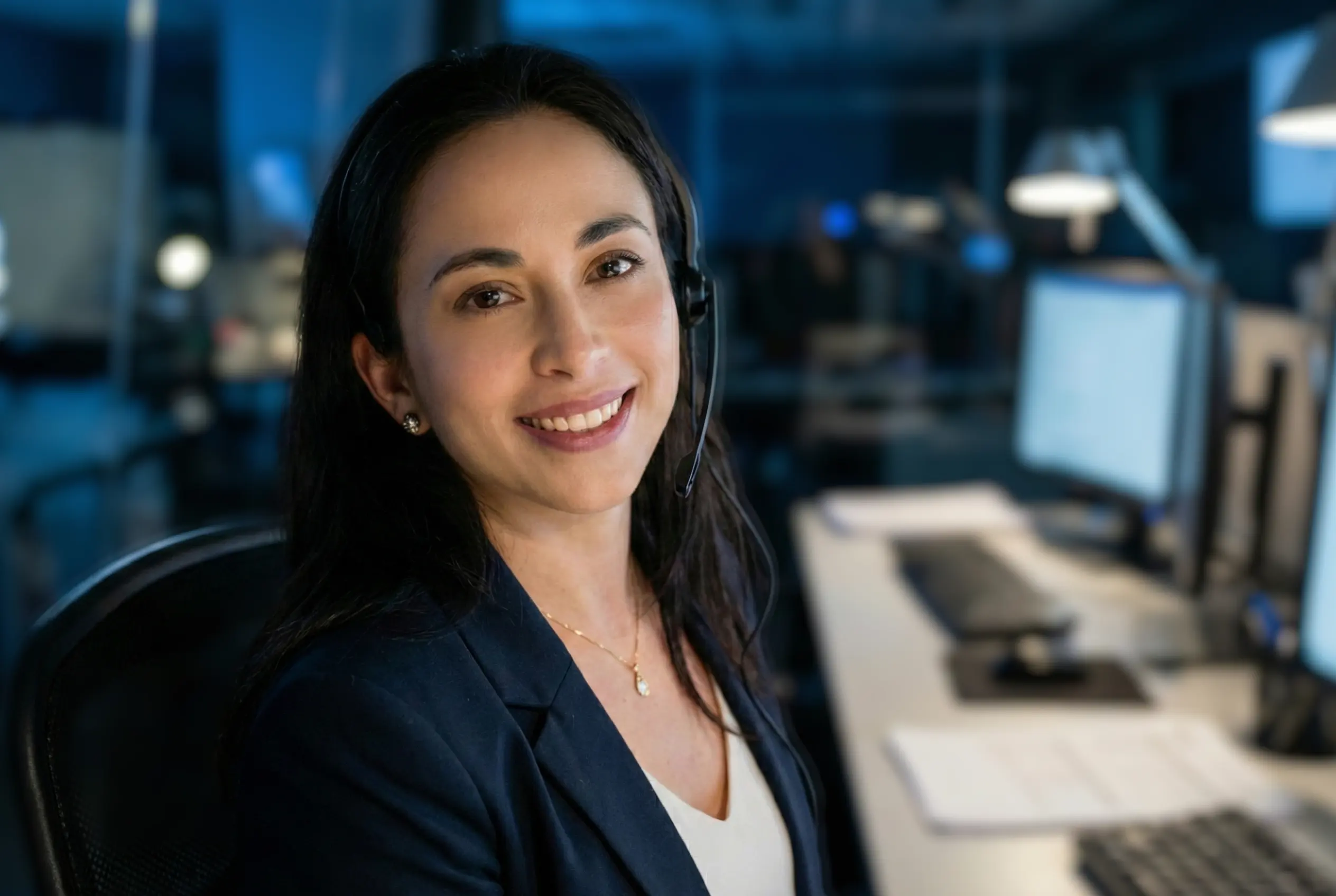 Smiling woman wearing headset and blazer sitting at a desk in a modern office with computer screens in the background.
