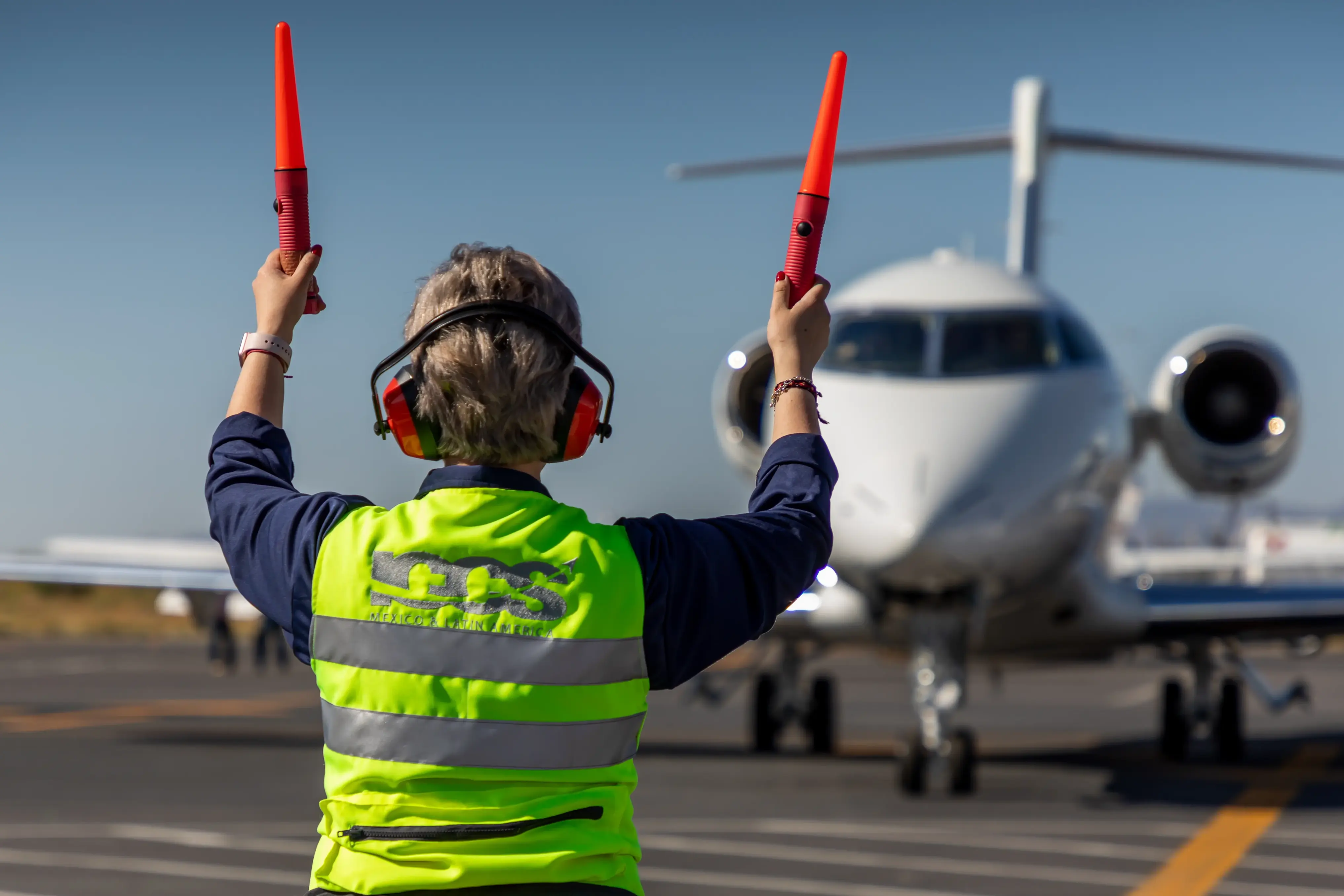 Airport ground staff in a yellow reflective vest directing a taxiing private jet using red marshalling wands.