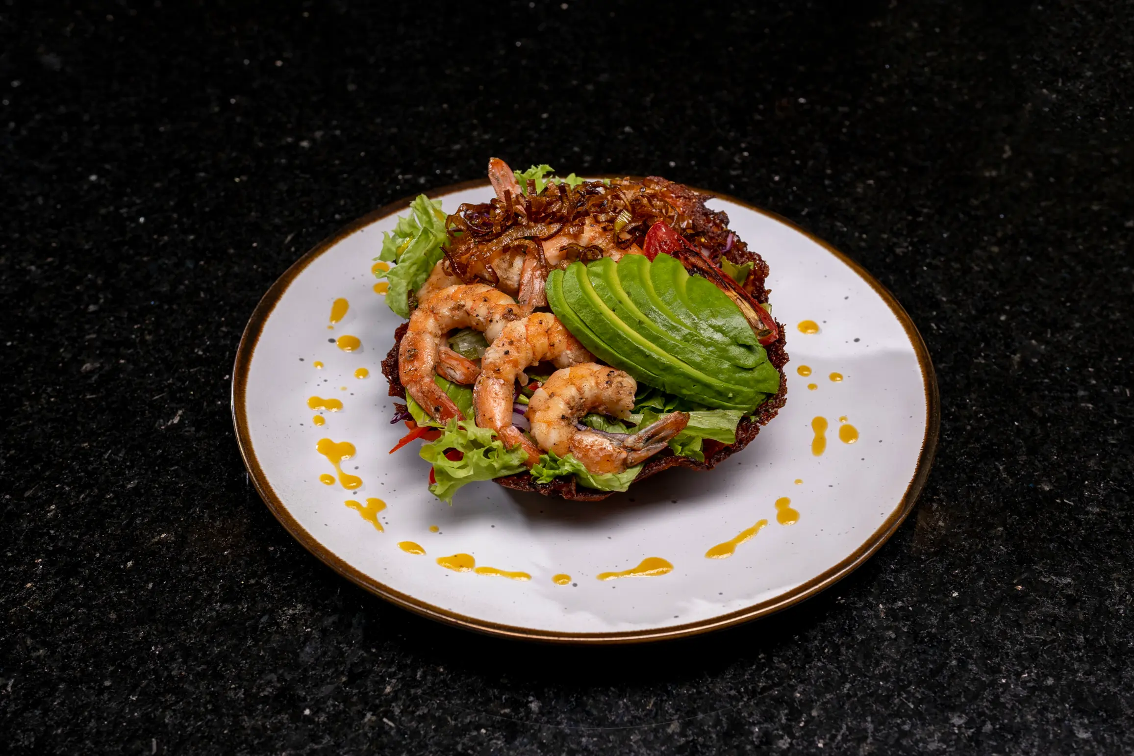 Grilled shrimp and sliced avocado salad on lettuce in a crispy bowl with drizzled dressing on a white plate.