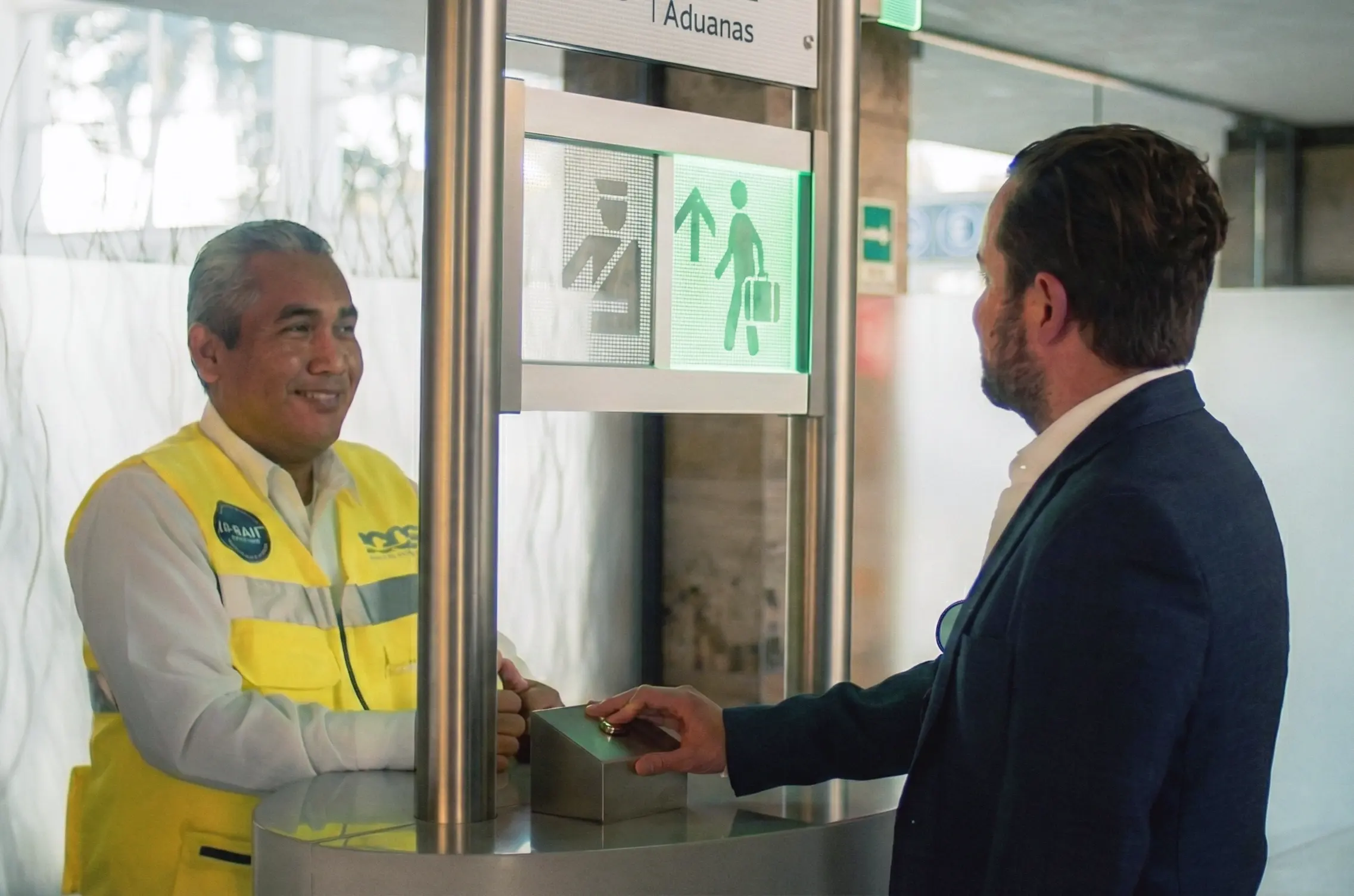 Customs officer in yellow vest smiling at a man in a suit who is presenting a passport at a customs checkpoint.
