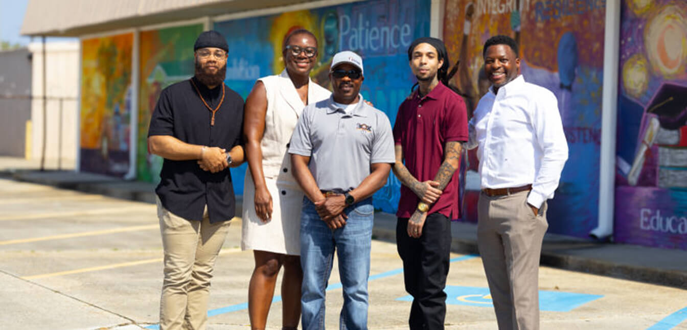 Five people smile and pose for a whole in front of a mural
