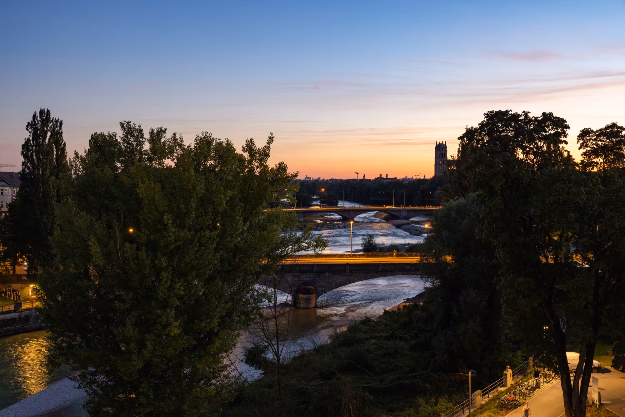 Ein Blick über Büsche und einen Fluss bei Abenddämmerung. Im Hintergrund sind zwei Türme zu sehen.