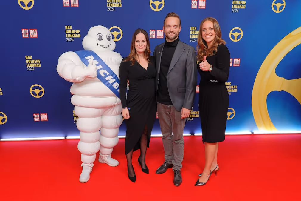 Drei Personen posieren mit dem Michelin-Männchen vor einer Sponsorenwand des goldenen Lenkrads.