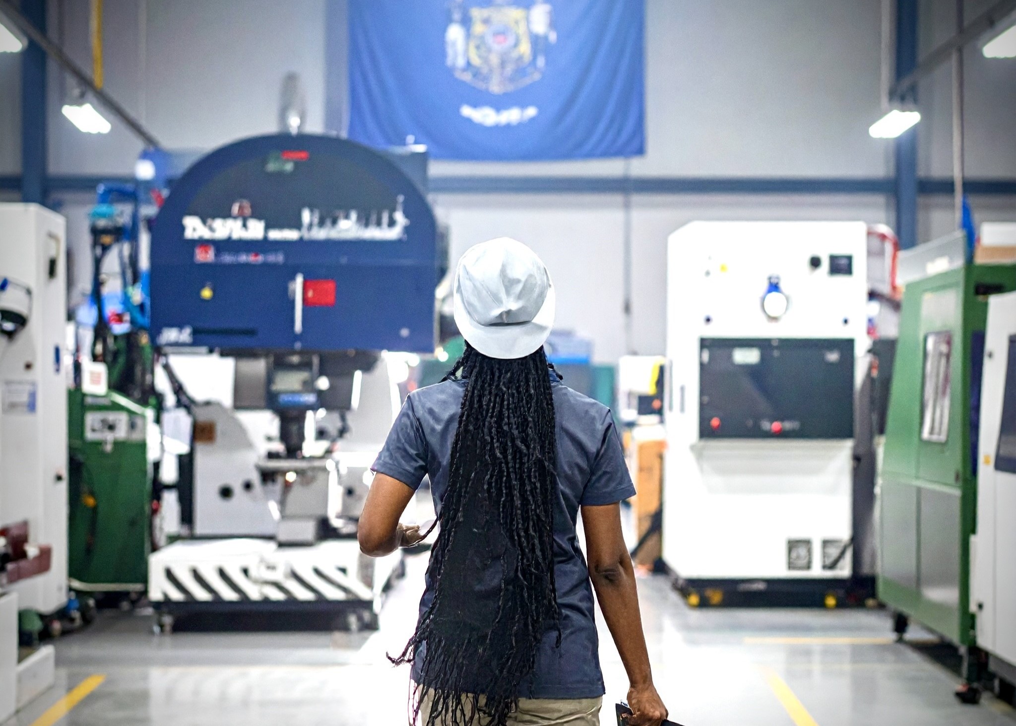 Person wearing a white hard hat walking in an industrial machine shop with large equipment.