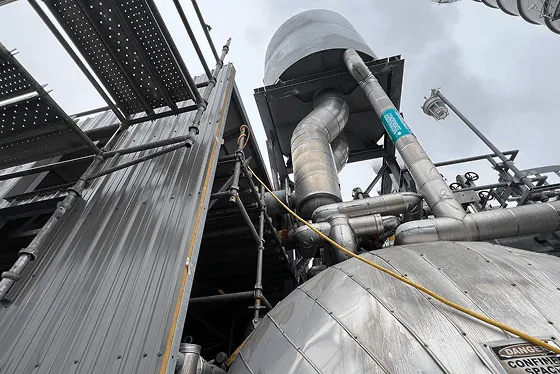 Industrial scene showing a large, gray metal structure with pipes and scaffolding. The cloudy sky adds a sense of industrial monotony and scale.