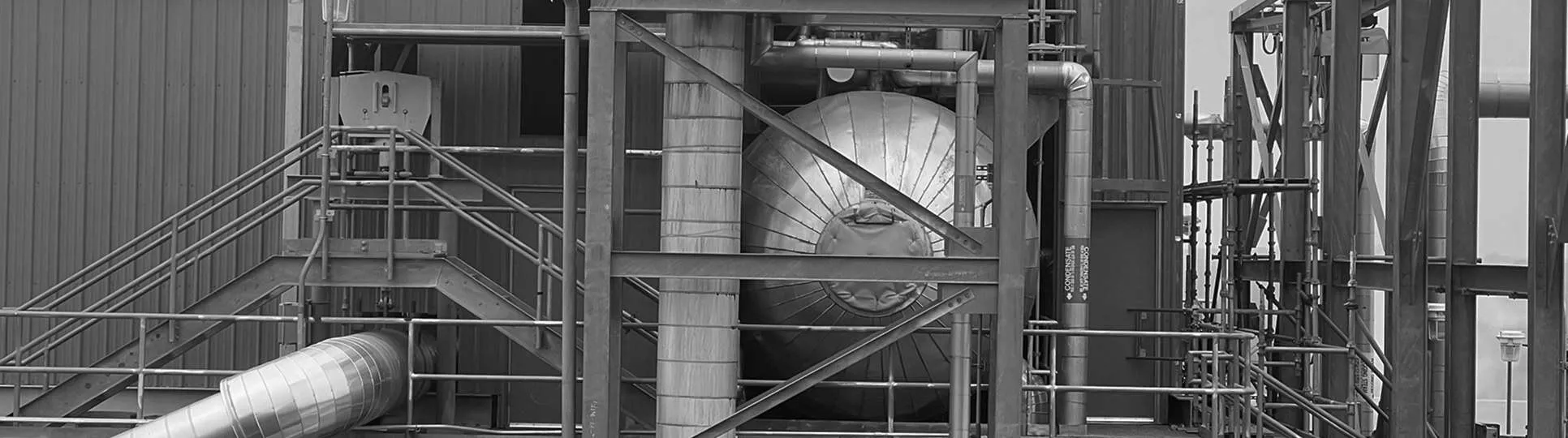 Industrial setting in black and white featuring metal beams, pipes, and a large cylindrical structure. The scene is complex and mechanical.