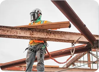 Construction worker wearing safety gear, including a helmet and harness, balances on steel beams while holding a wooden plank at a building site.