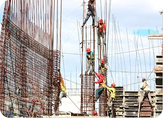 Group of construction workers with helmets scale steel beams at a construction site, coordinating teamwork under a bright, partly cloudy sky.