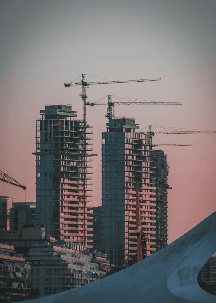 Tall construction cranes surround modern high-rise buildings during a serene sunset, casting a pink and blue hue over the urban skyline.