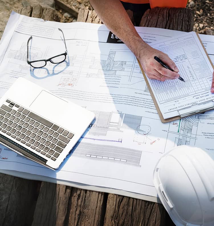 A person reviews architectural blueprints on a wooden table, with a laptop, glasses, and hard hat nearby, conveying focus and analytical work.