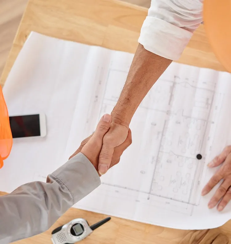 Two construction professionals shake hands over architectural plans on a table. One wears an orange safety vest. A phone and two-way radios are nearby, conveying cooperation.