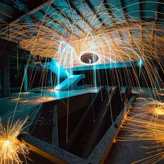 A dimly lit industrial space is illuminated by swirling orange and blue light trails above an escalator, creating a dynamic and energetic atmosphere.