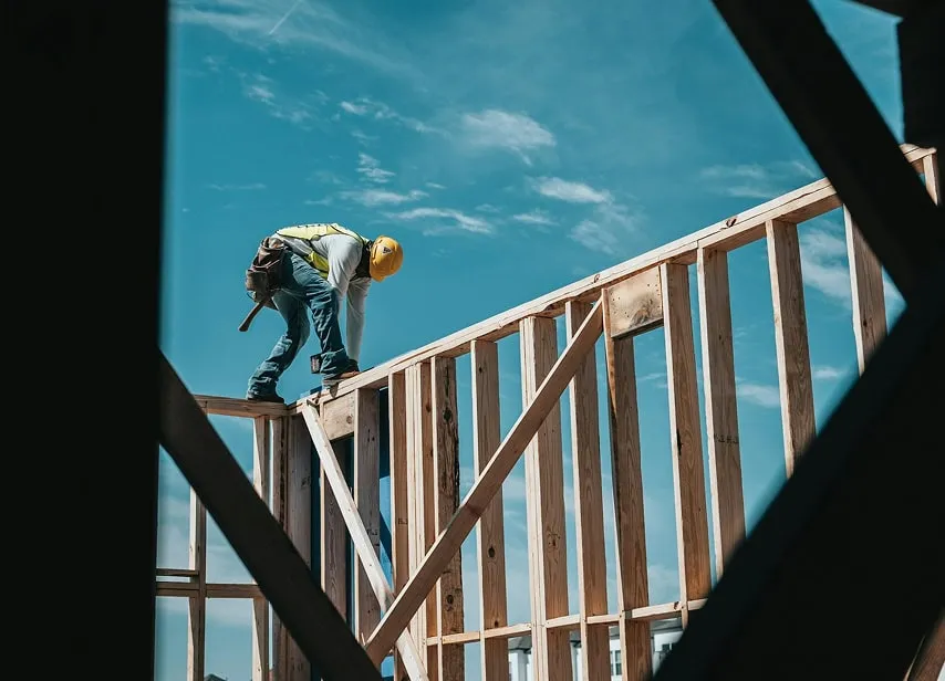 A construction worker in a yellow hard hat and vest balances on a wooden frame against a clear blue sky. The scene conveys focus and caution.