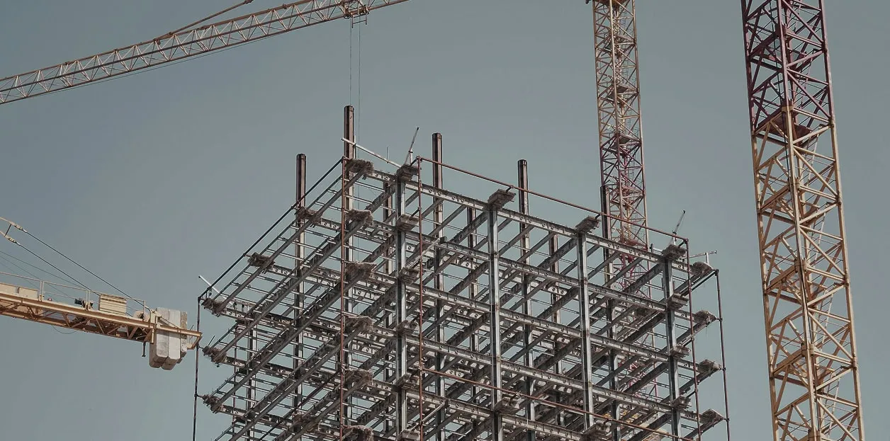 Steel framework of a building under construction with two tall cranes against a clear blue sky, conveying a sense of progress and industrial strength.