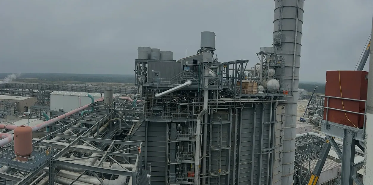A sprawling industrial facility with large metallic structures and tall chimneys under a gray, overcast sky, conveying a sense of scale and complexity.
