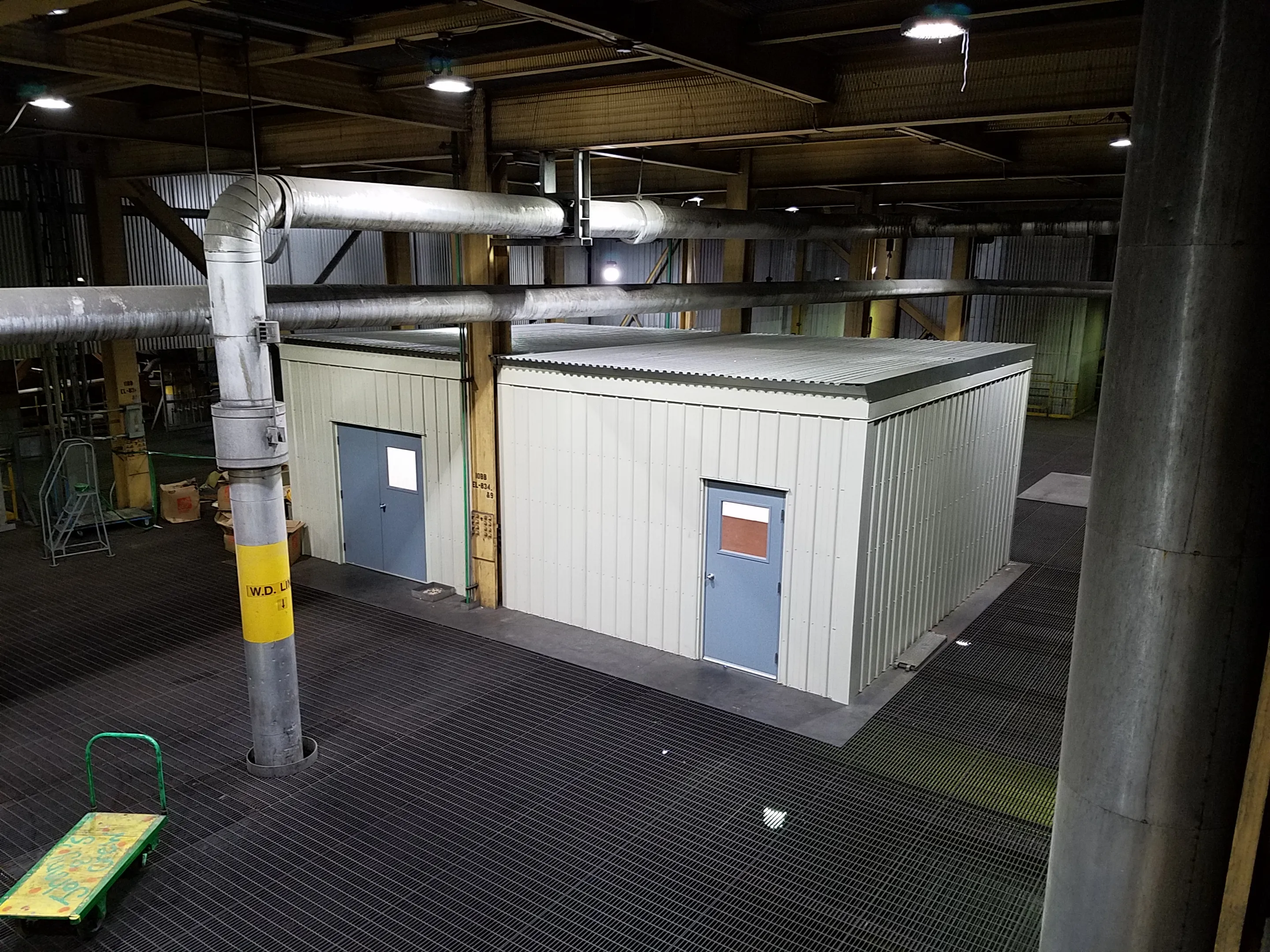 Interior of an industrial facility showing a small prefabricated metal building with two blue doors and overhead ductwork.