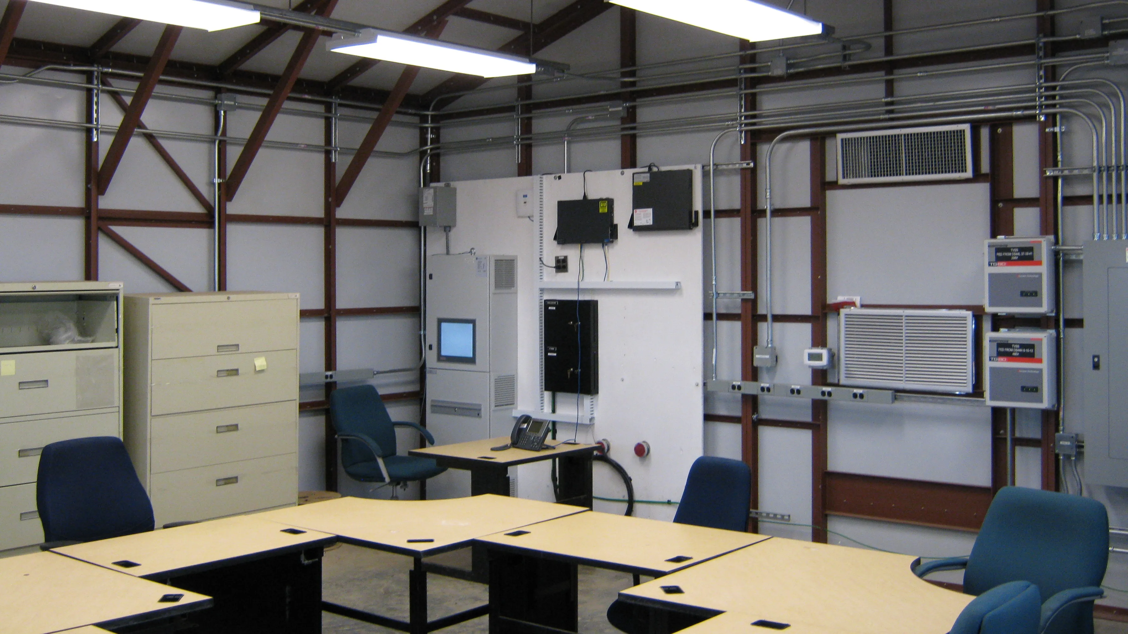Industrial office room with beige filing cabinets, blue office chairs, connected tables, and wall-mounted control panels with wiring.