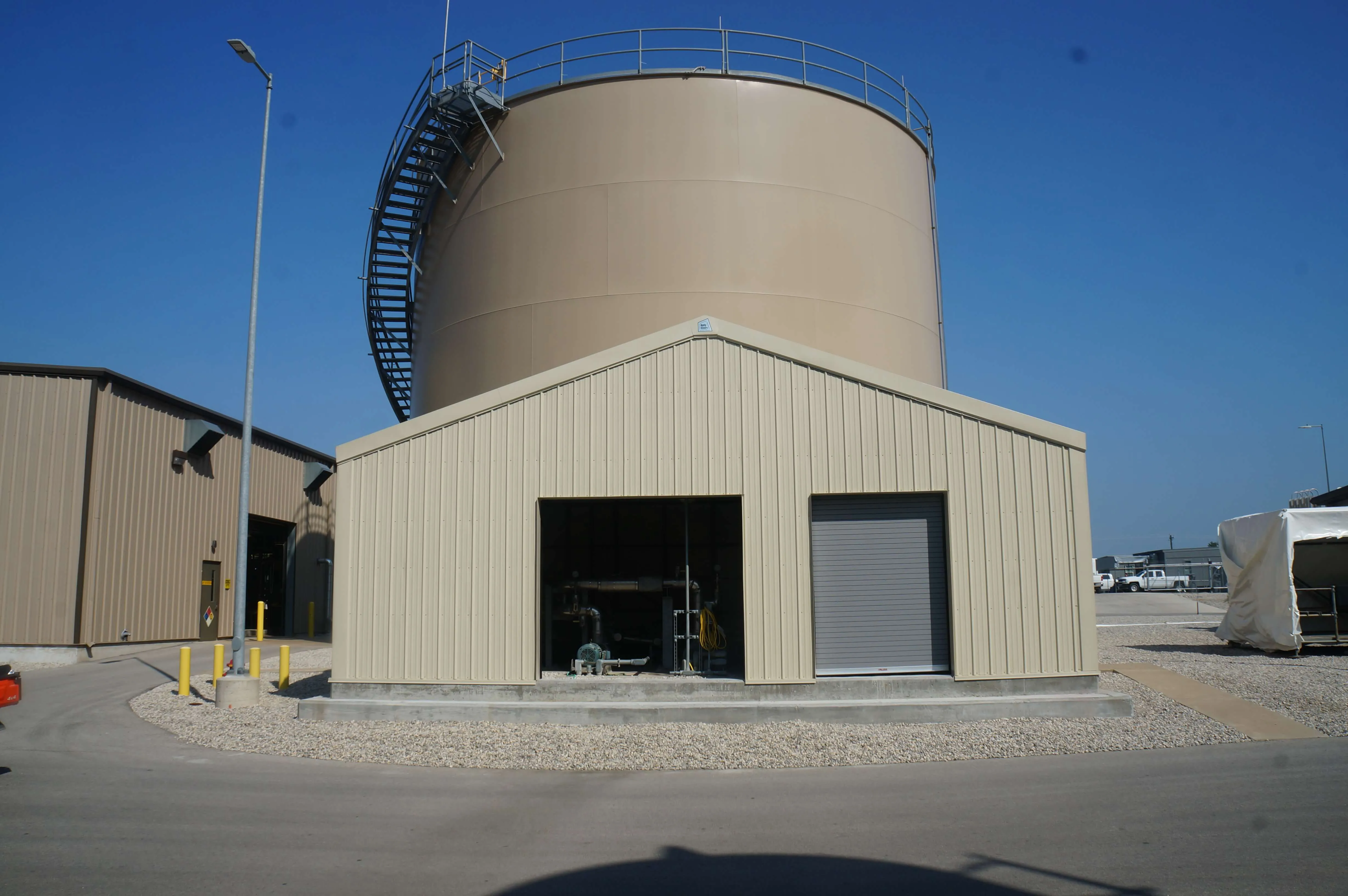 Large beige industrial storage tank with spiral exterior staircase behind a small beige building with an open section showing pipes and a closed roll-up door.
