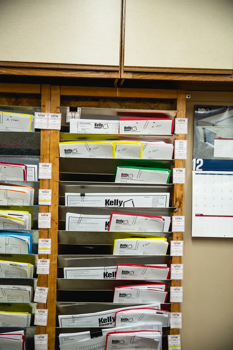Wooden rack filled with organized, color-coded folders labeled Kelly Klosure and assorted project codes next to a wall calendar.