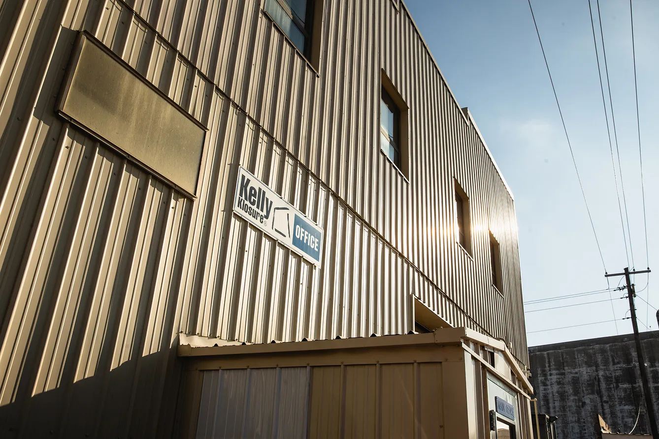 Exterior of a beige metal-clad commercial building with a sign that reads 'Kelly Klosure OFFICE' and sunlight reflecting off the windows.