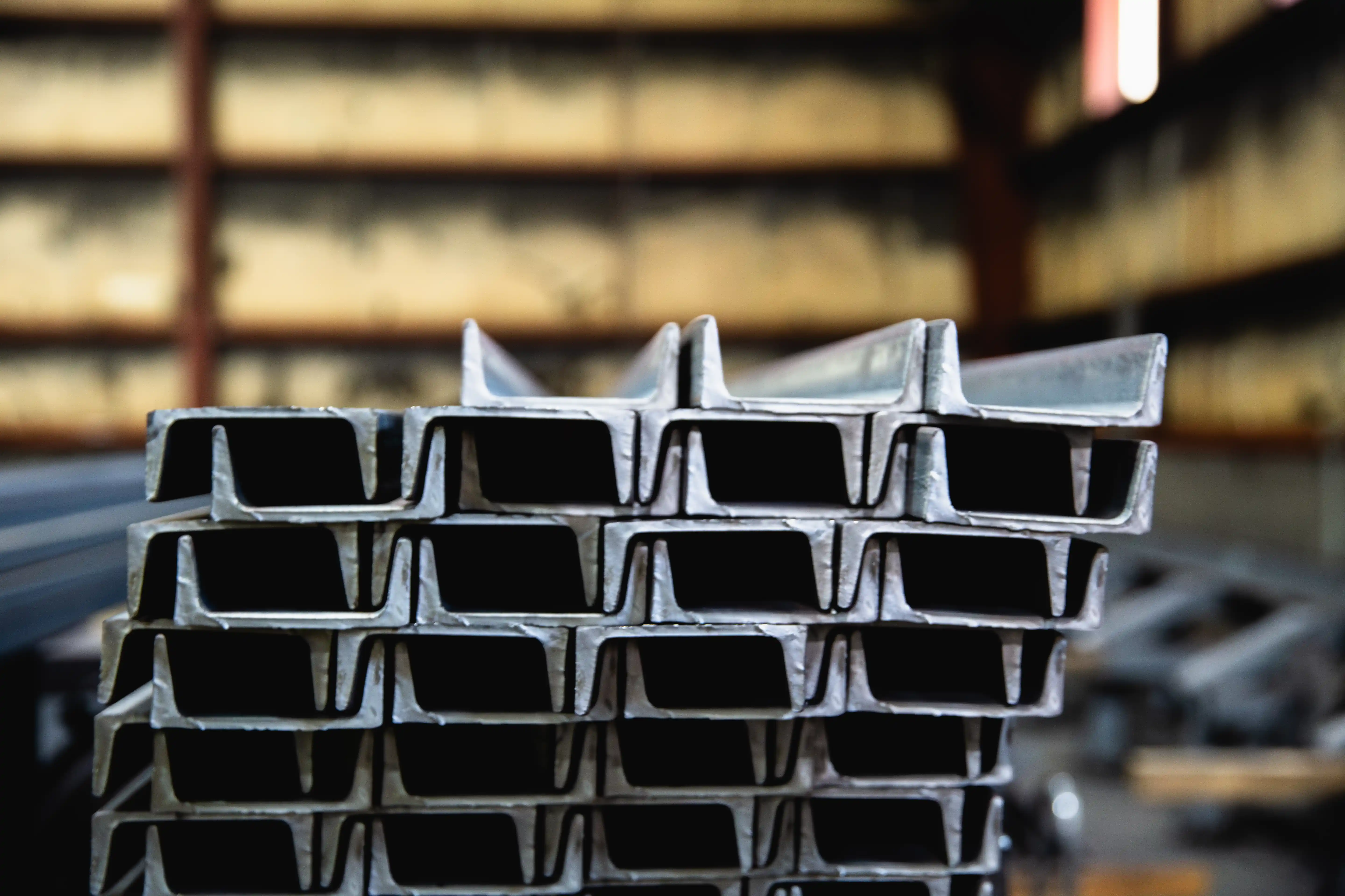 Stack of metal I-beams inside a warehouse with blurred industrial background.