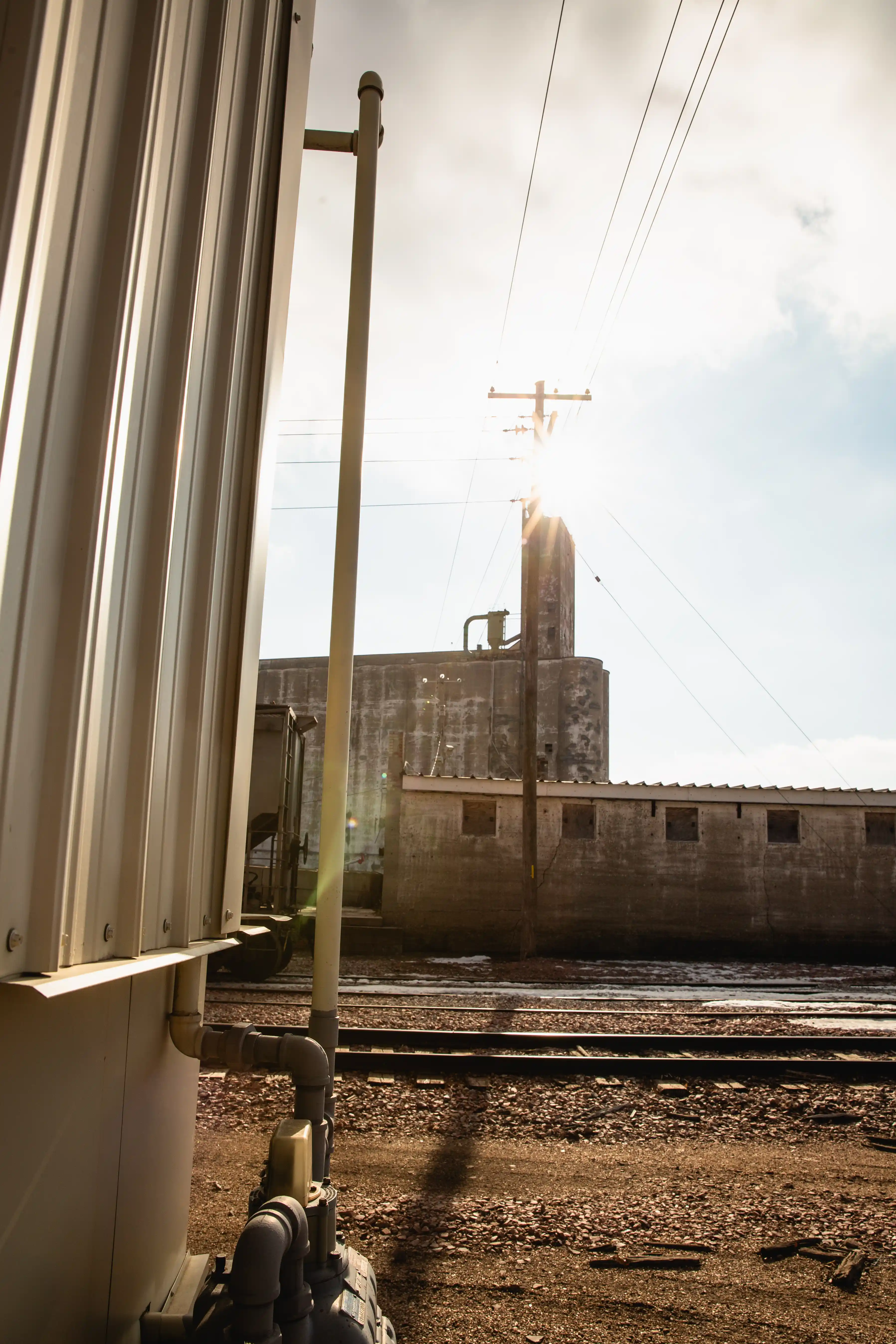 Sun shining behind a utility pole near railroad tracks and industrial buildings with pipes in foreground.