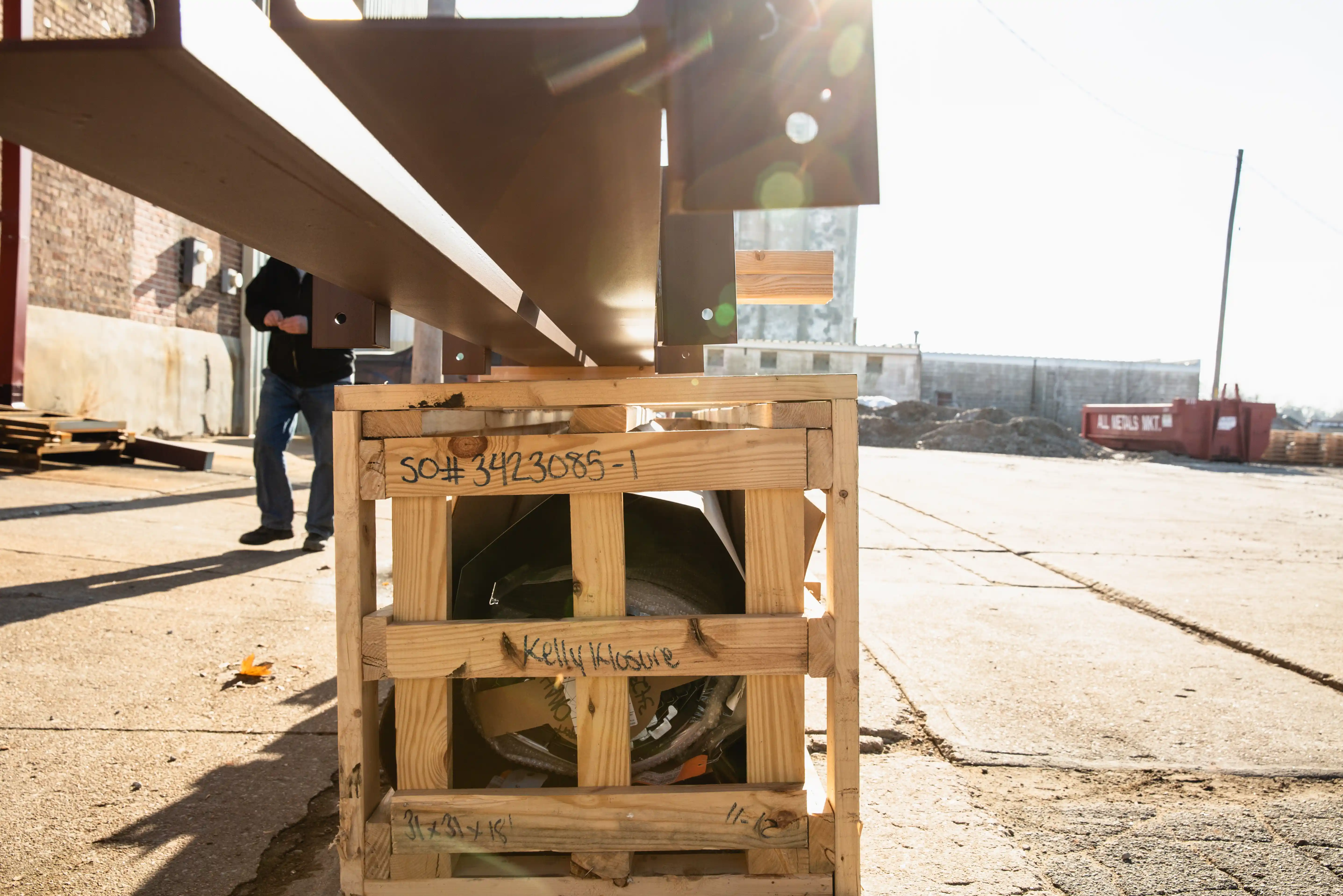 Wooden crate holding metal parts on a sunny outdoor industrial site with a person walking nearby.