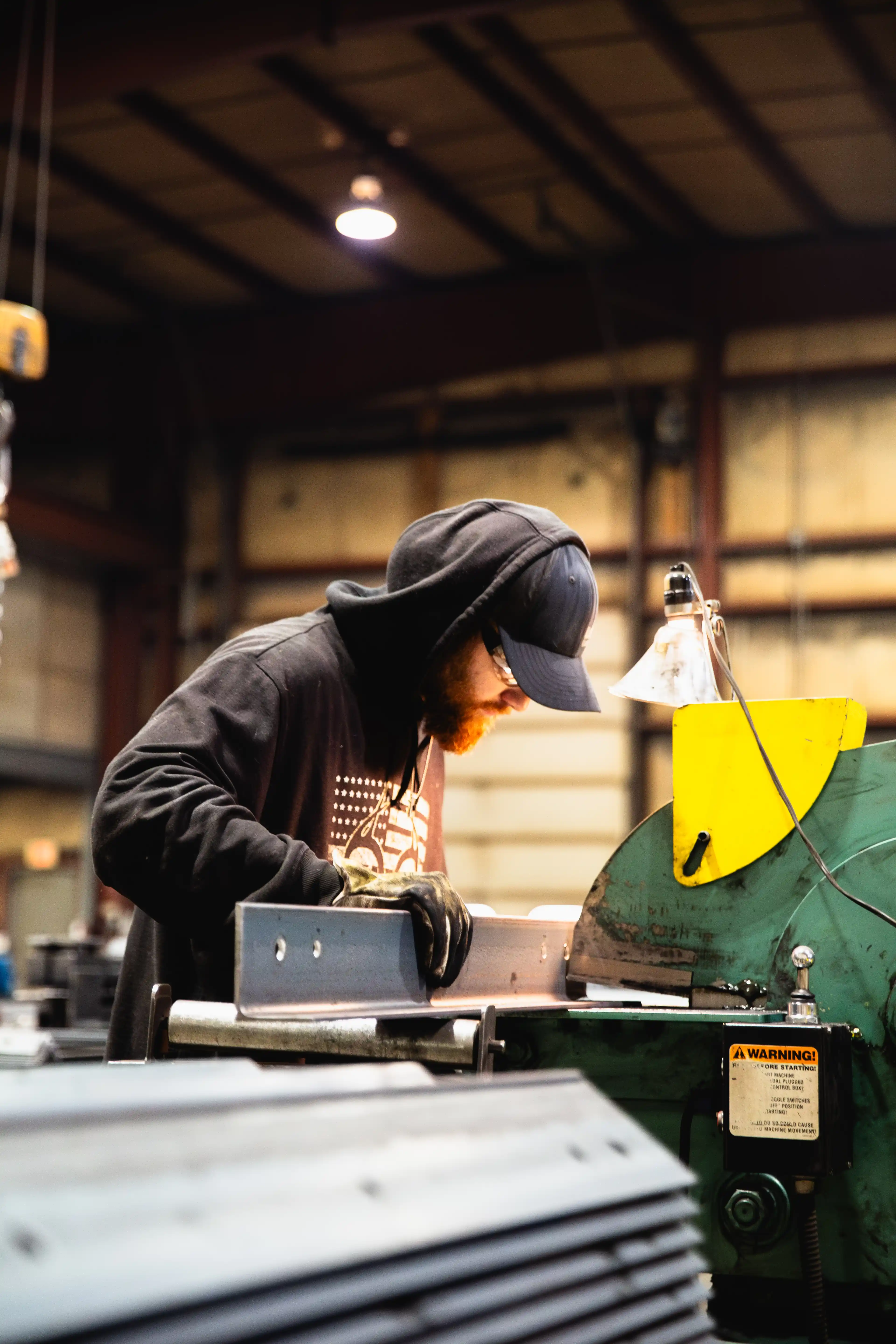 Worker wearing gloves and hoodie inspecting metal piece in an industrial workshop.