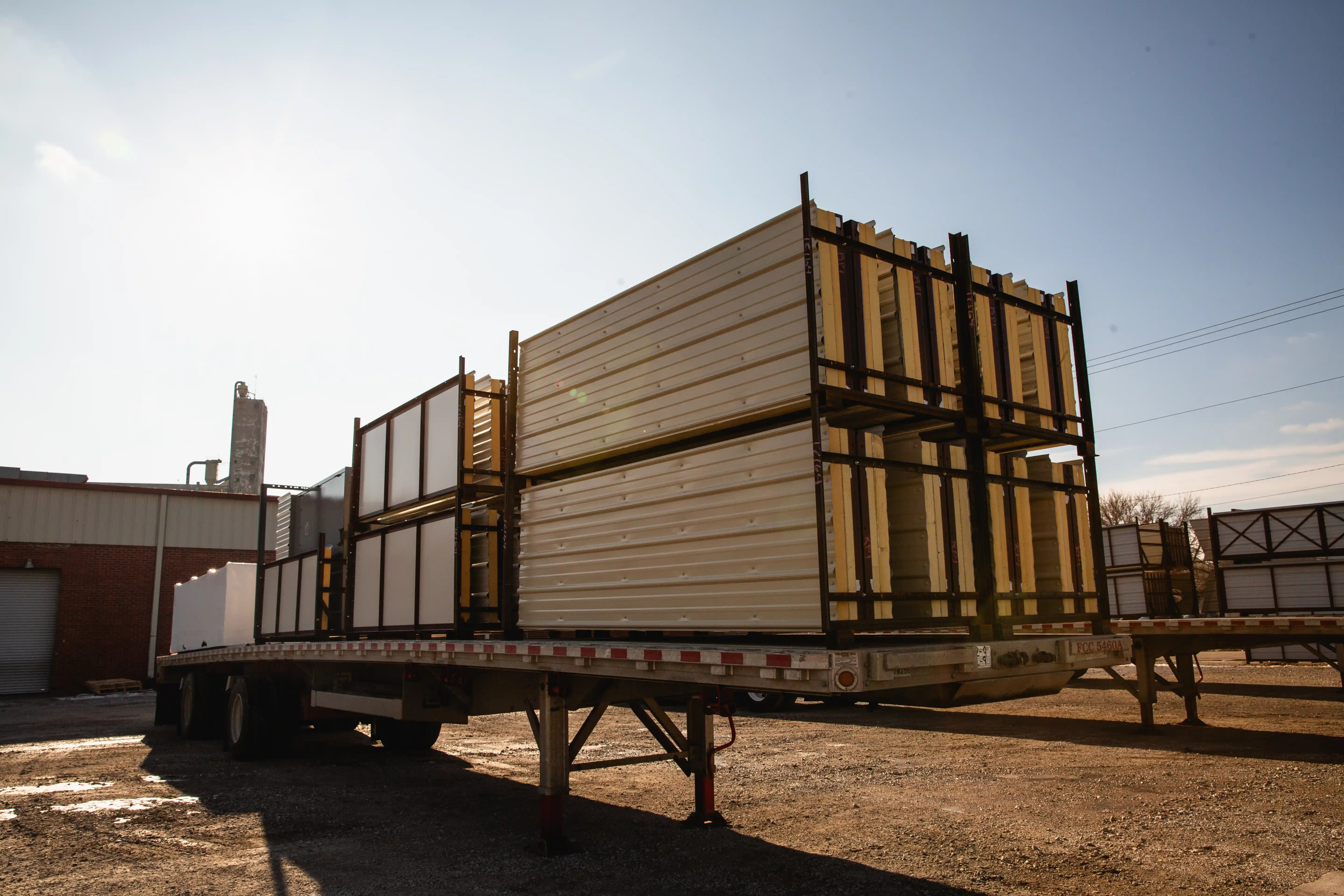 Flatbed trailer loaded with large metal-framed panels in an industrial outdoor yard at sunset.